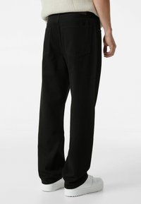 Pantalones de mezclilla negros con un corte recto, que cuentan con dos bolsillos traseros y una textura suave, usados con zapatillas blancas.