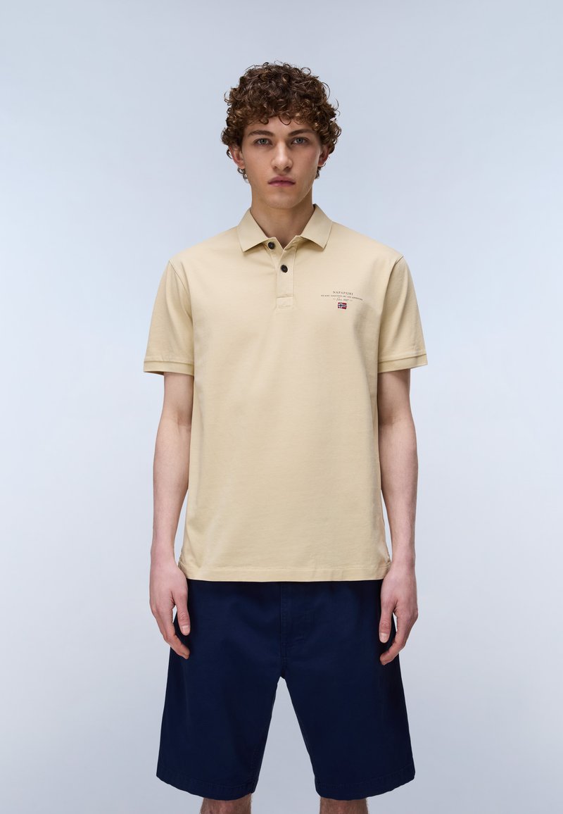 Jeune homme aux cheveux bouclés portant un polo beige avec un petit logo et un short bleu marine, debout devant un fond clair uni.