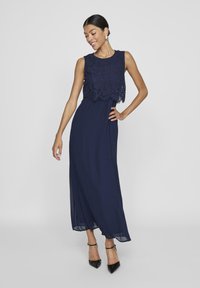 Mörkblå aftonklänning med ärmlös spetsöverdel och flödande kjol. Har en figursydd bodice och ett transparent lager vid fållen.