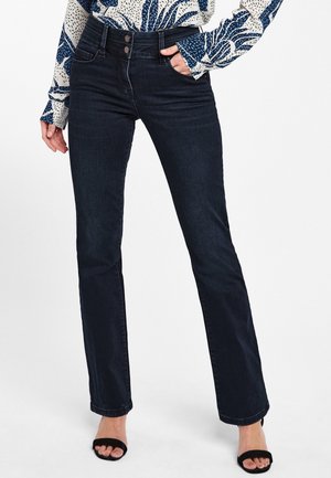 Femme portant un jean droit bleu foncé et un chemisier à motifs, une main dans la poche et des talons ouverts noirs.