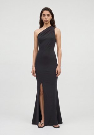 Un mannequin se tient devant un fond clair uni, portant une robe noire longue jusqu’au sol, à une épaule dénudée, avec une fente jusqu’à la cuisse et des sandales.