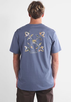 T-shirt en coton bleu avec un motif floral dans des couleurs neutres au dos, contenant la marque "Oxbow" imprimée sous le graphique.