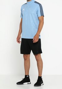 Man som bär en ljusblå Adidas kortärmad sporttop, svarta shorts, svarta Adidas strumpor och svarta löparskor som står på vit golv.