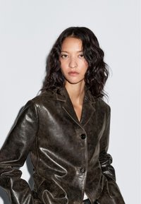 Vrouw met golvend donker haar, gekleed in een donkerbruine leren jas, staande tegen een effen lichte achtergrond, met een neutrale uitdrukking naar voren kijkend.