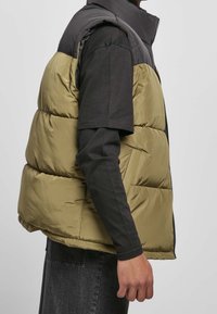 Pufferväst med svart överdel och olivgrön nedre del. Har tjock vaddering, hög krage och dragkedja framtill.