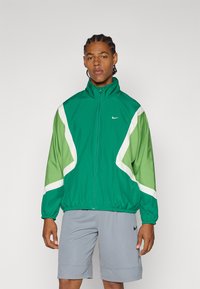 Žalias Nike vėjara su baltais ir tamsiai žaliais akcentais. Yra aukštas apykaklė, užtrauktuko uždarymas ir elastiniai rankogaliai, derinami su pilkais šortais.
