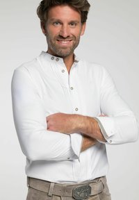Witte langemouwen shirt met een hoge kraag, decoratieve knopen en subtiele piping-details; gedragen met een grijze suede riem met een metalen gesp.