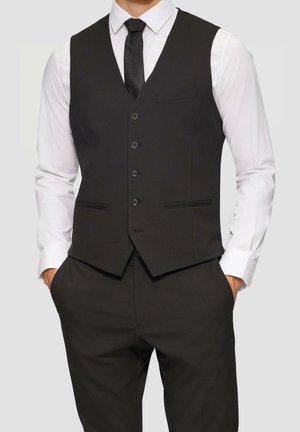 Mann trägt schwarze, geknöpfte Weste, schwarze Krawatte, weißes Hemd mit langen Ärmeln und passende schwarze Hose, Hände in den Taschen, Oberkörper sichtbar.