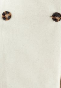 Kreemikas puuvillakangas sujuva tekstuuriga, kus on kaks ümmargust kilpkonnakujulist nööp. Lihtne, puhas disain koos peene mustriga.