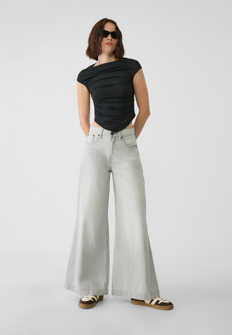 Haut noir ajusté avec une texture froncée, associé à un jean large gris clair. Le modèle porte des lunettes de soleil sombres et des baskets marron avec des rayures blanches.
