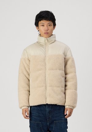 Veste beige matelassée avec un devant en polaire texturée douce, dos en tissu lisse, col montant et fermeture éclair. Coupe décontractée, poignets élastiques.