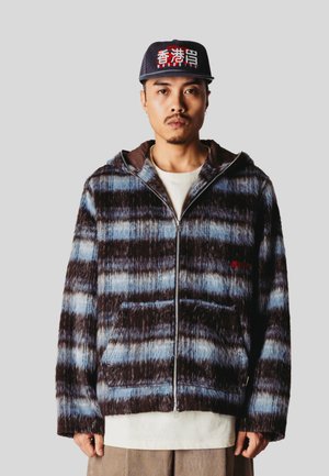 Homme portant une veste zippée à carreaux bleus et marron, un pantalon beige et une casquette noire avec un texte rouge et blanc sur un fond gris uni.
