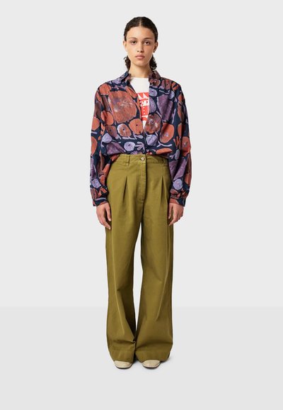 Donna in piedi con una camicia a bottoni viola e arancione, una maglietta bianca con grafica, pantaloni larghi color oliva e scarpe beige, su uno sfondo grigio.