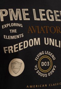 Camiseta de algodón negra con varios textos impresos en crema y marrón. Presenta logotipos y frases como "Aviadores" y "Leyenda Voladora DC-3."