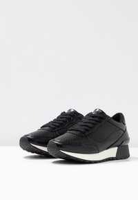 Paire de baskets en cuir noir avec côtés texturés, lacets noirs et semelle épaisse blanche et noire sur surface blanche.