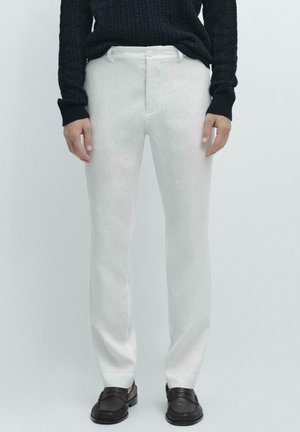 Uomo che indossa pantaloni bianchi a taglio dritto, maglione nero lavorato a maglia e mocassini in pelle nera, in piedi contro uno sfondo semplice.