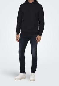 Sudadera negra de mezcla de algodón, con un bolsillo tipo canguro y cordones ajustables. Combinada con jeans de mezclilla oscuros y zapatillas blancas.