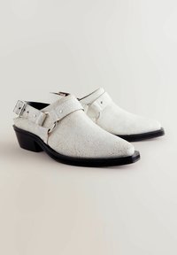 Valkoiset halkeillut nahasta valmistetut slip-on-kengät, joissa on matala musta korko, säädettävä nilkkastrappi ja metalliset korostukset. Teksturoitu pinta lisää visuaalista kiinnostavuutta.