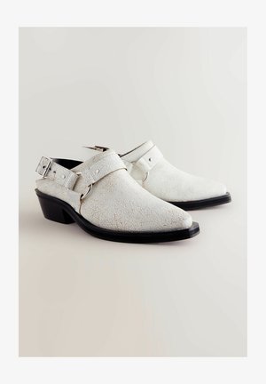 Valged pragunenud nahast libisemisega kingad madala musta kontsaga, reguleeritava hüljangiga ja metallaktsentidega. Tekstureeritud pind lisab visuaalset huvi.