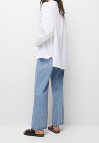 Lockere, weiße Leinenbluse mit langen Ärmeln, kombiniert mit hellblauen, weit geschnittenen Jeans und schwarzen Sandalen. Entspannte Silhouette, minimalistisches Design.