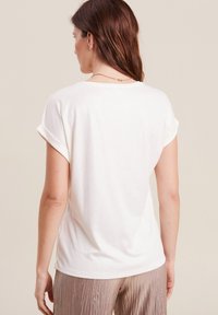 T-shirt bianco a maniche corte realizzato in tessuto morbido e leggero. Presenta maniche arrotolate e una vestibilità rilassata. La vista posteriore mostra una texture liscia e una leggera lucentezza.