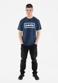 Navy blue T-shirt with “oasis” logo in white and light blue within a rectangular frame; paired with black trousers and dark shoes.