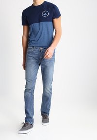 Camiseta de color bloqueado en azul marino y azul con un logo circular en el pecho, combinada con jeans de un azul claro y zapatillas grises con suelas blancas.