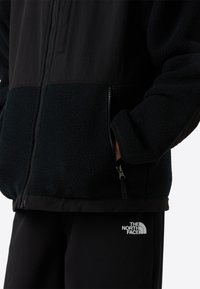 Flis jakna u crnoj boji s glatkim gornjim dijelom i teksturiranim donjim dijelom. Ima džepove sa zatvaračima i logotip na donjoj desnoj strani.