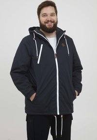 Ung mann med skjegg iført en svart hettejakke med hvit glidelås og snorer, med hendene i lommene, smilende mot en ensfarget bakgrunn.