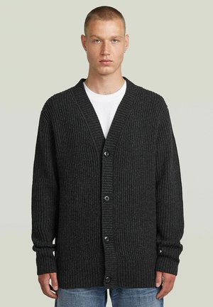 Jeune homme aux cheveux courts portant un cardigan côtelé gris foncé boutonné sur une chemise blanche et un jean bleu sur un fond uni.