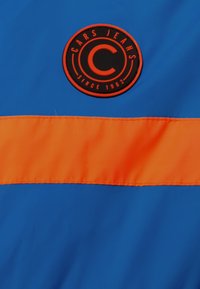 Modrá tkanina s oranžovým horizontálním pruhem a kulatou černou a oranžovou nášivkou s nápisem "CARS JEANS SINCE 1982." Hladký povrch.