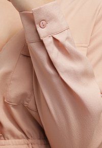 Ljusrosa blusärm i mjukt, texturerat tyg med en liten knappdetalj vid manschetten. Har subtila veck vid axeln.