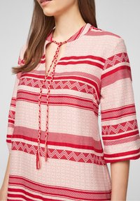 Roze tuniek met patroon en rode horizontale strepen, met een ritssluiting en een gevlochten strik. Korte mouwen met geometrische ontwerpen.