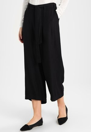 Kobieta nosząca czarne, szerokie, skrócone spodnie z materiałowym paskiem, połączone z czarnymi balerinami i białą, teksturowaną bluzką z długim rękawem.