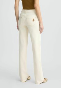 Jean évasé crème clair avec poches arrière et broderie logo dorée. Texture en denim lisse et design de taille standard.