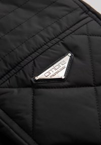 Sort quiltet stof med et metallic sølv trekantet logo, der står "ombre menswear." Teksturen er glat med detaljeret syning.