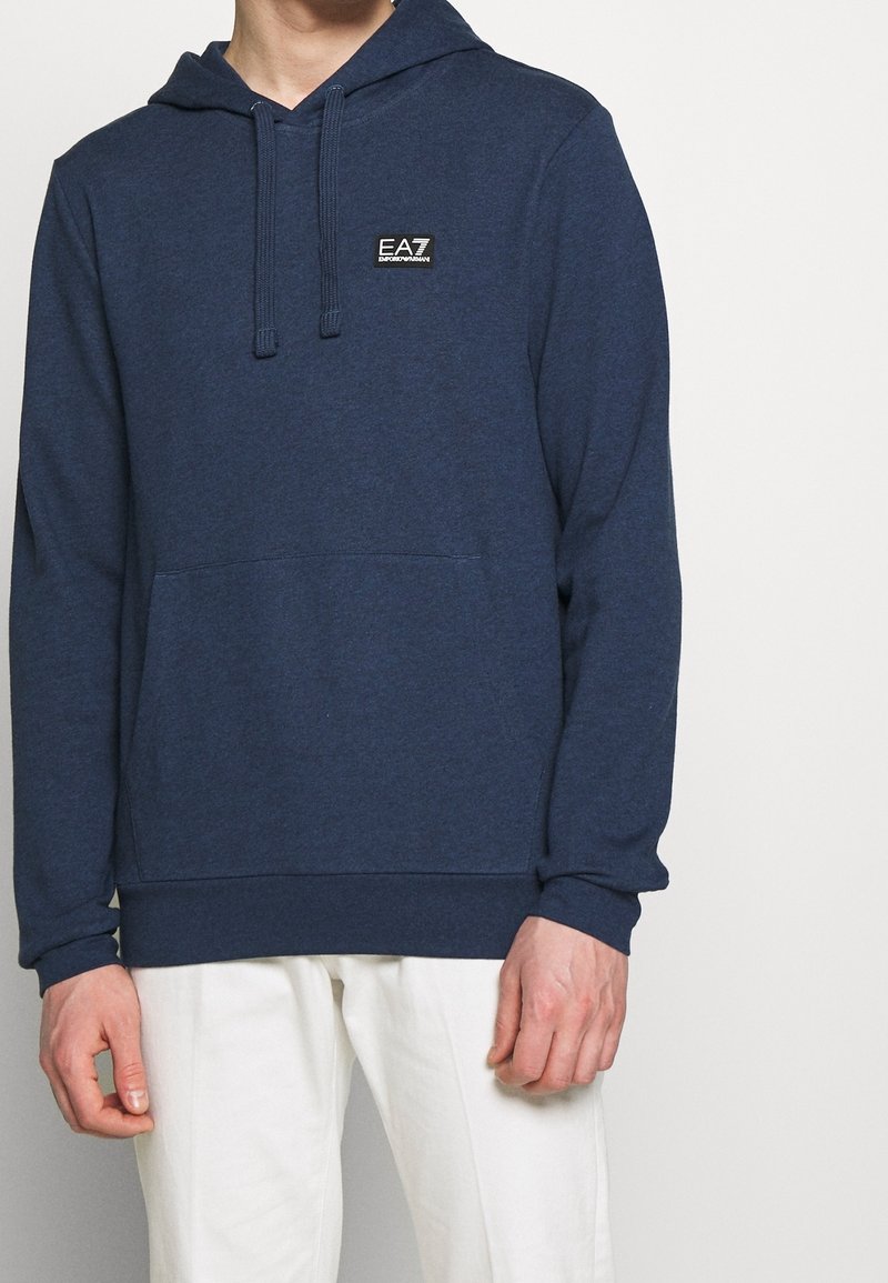 Homme portant un sweat à capuche bleu marine EA7 Emporio Armani avec poche avant et pantalon blanc sur fond uni.