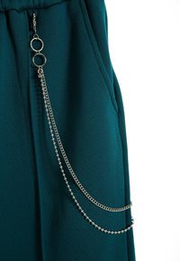 Pantalones de chándal en color verde azulado con cintura ajustable mediante cordón, cuentan con un detalle de cadena plateada en el lado, compuesto por dos capas con cuentas y eslabones metálicos.