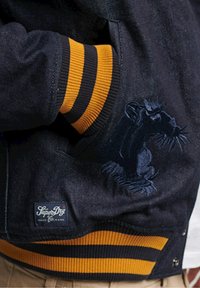 Veste en denim foncé avec une broderie de rat sur la poche gauche, poignets côtelés avec des rayures noires et jaunes, étiquette de marque à l'ourlet.