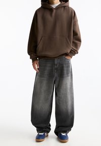 Ruskea ylikokoinen huppari, jossa on etutasku, yhdistettynä väljiin harmaan sävyn liukuvärjättyihin farkkuihin ja sinisiin lenkkareihin, joissa on valkoisia yksityiskohtia.