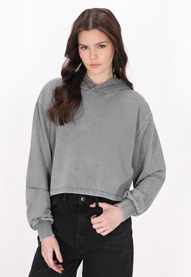 Felpa grigia accorciata con una texture morbida e cappuccio con lacci. Maniche lunghe, leggermente svasate, con polsini a costine. Indossata sopra jeans neri.