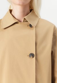 Trench coat bej cu un design drept, guler mare și două butoane în față. Țesătura pare netedă și ușoară.
