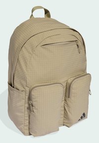 Mochila beige claro con un patrón de cuadrícula texturizado, compartimento principal con cremallera, dos bolsillos frontales y un asa superior. Presenta detalles de logo en negro.