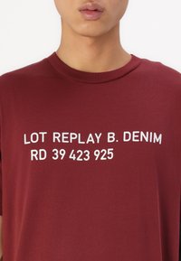 Bordová bavlněná trička s krátkým rukávem s bílým nápisem: "LOT REPLAY B. DENIM RD 39 423 925" výrazně tištěným na přední straně.
