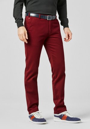 Mann trägt rote Slim-Fit-Hose, dunklen Gürtel, schwarzen langärmligen Oberteil und blau-weiße Sneakers mit orangen Streifen vor einfarbigem Hintergrund.