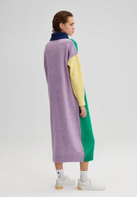 Robe tricotée longue avec un mélange de sections en violet, vert, jaune et marine ; dispose d'un col haut et d'un ourlet côtelé. Portée avec des baskets blanches.