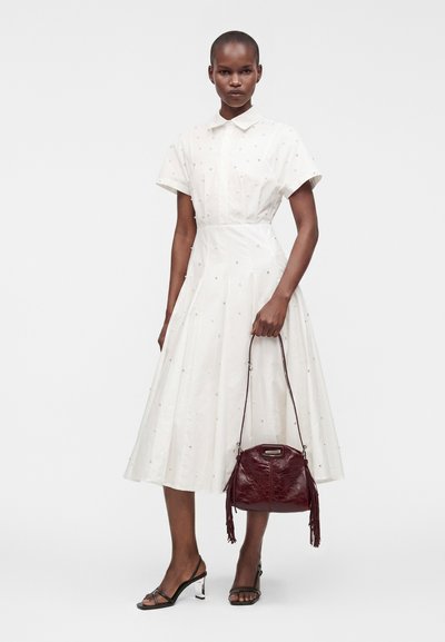 Femme portant une robe midi blanche à manches courtes avec de petits ornements, tenant un sac à main rouge foncé et portant des talons ouverts noirs.
