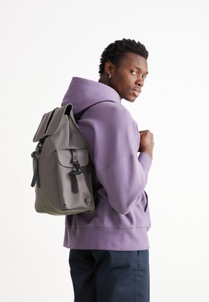 Szary plecak z klapą i przednią kieszenią, posiadający czarne okucia i paski; noszony na fioletowej bluzie z kapturem.
