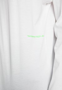 Weißes Baumwollhemd mit langen Ärmeln, das auf der unteren Vorderseite grünen Text mit der Aufschrift "'Not Ordinary' REPLAY" zeigt. Weicher Stoff, minimalistisches Design.