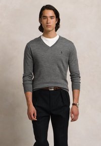Homem vestindo um suéter cinza em V sobre uma camisa branca, calças pretas com um cinto castanho e um relógio de pulso, em pé contra um fundo neutro.